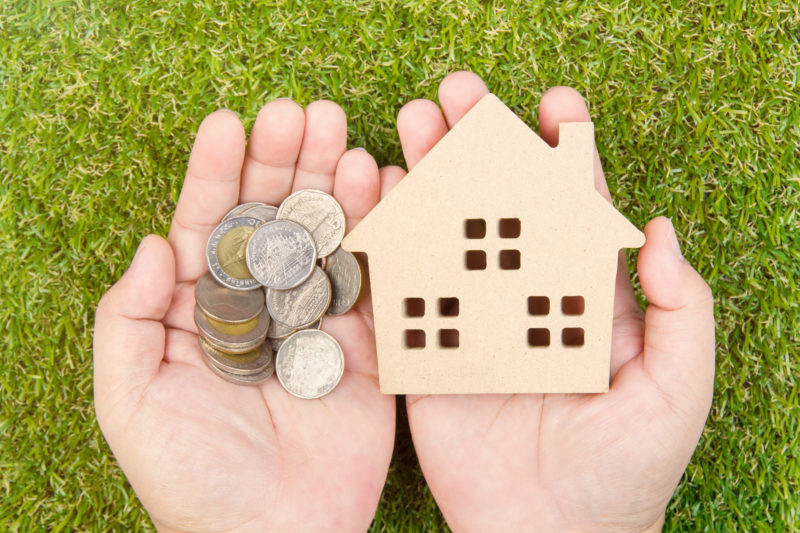 Mortgage concept image. A pair of hands hold a small wooden house and a pile of coins, set against a background of grass.