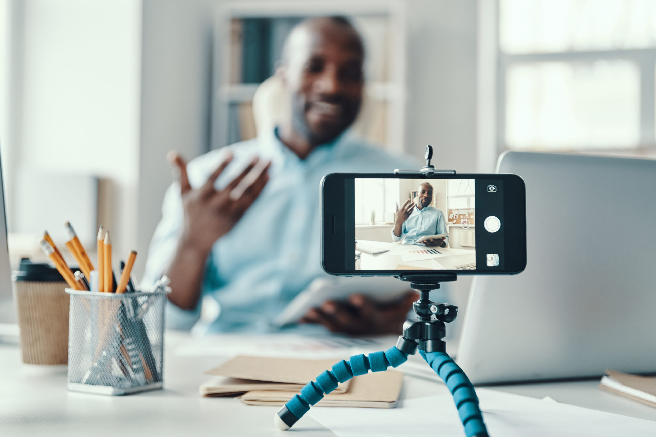 man recording a video on a smartphone placed on a flexible tripod, sitting at a desk with office supplies, a laptop, and papers in a bright, modern workspace